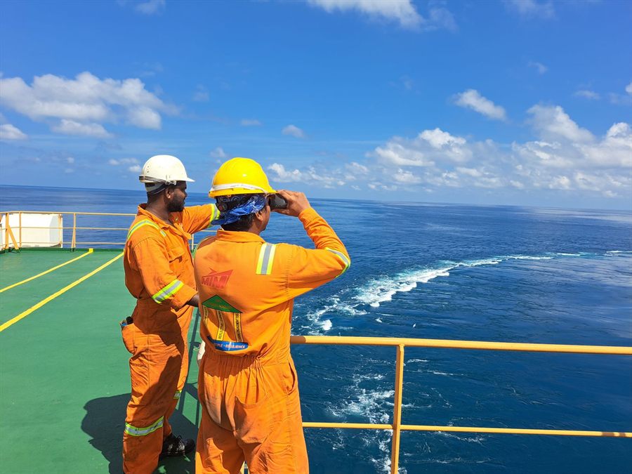 Two seafarers in orange overalls and hard hats watching the sea from a ship&rsquo;s deck